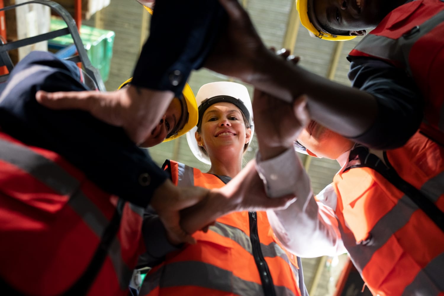 A diverse group of construction workers wearing safety gear form a circle, hands stacked in teamwork. They look up with smiles, conveying unity and collaboration.