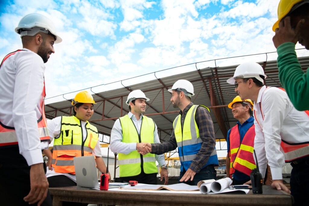 A diverse group of construction workers wearing safety vests and hard hats gather around a table with blueprints, two men shaking hands, conveying teamwork.