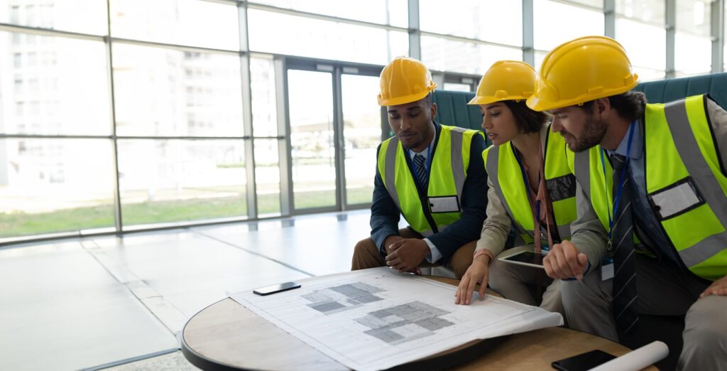 Three construction professionals in hard hats and reflective vests review architectural plans on a table in a bright, modern office setting.